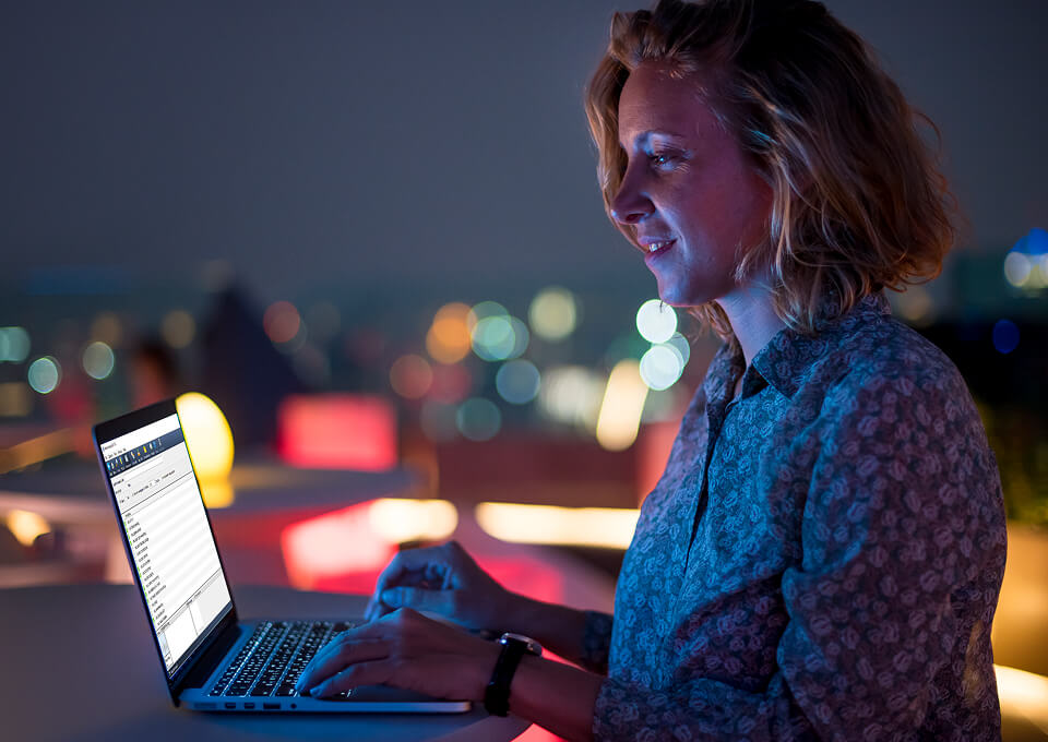 A woman is sitting outside on a balcony and using a laptop. She is smiling at the Newshosting Newsreader newsgroup search.