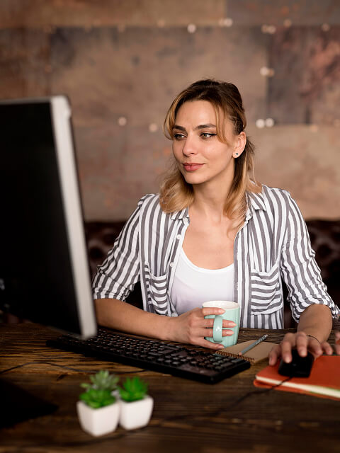 A woman in a black and white striped shirt is sitting at a desk holding a coffee cup and working on a desktop computer.