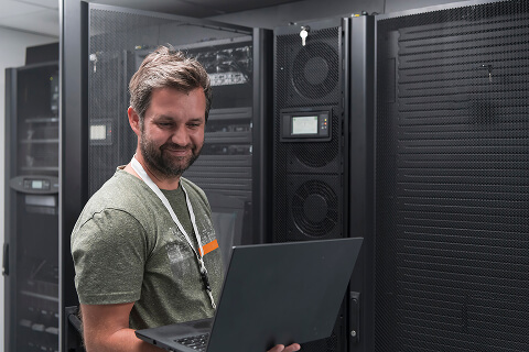 A male technician is working on maintaining the Newshosting network. He is holding a laptop on his right forearm and smiling.
