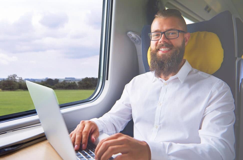 A man with a beard and black glasses is on a train. He is typing on a laptop and looking at the camera, smiling.
