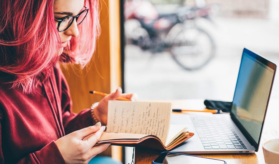 A woman with pink hair and a red sweater is browsing Usenet on her laptop and is taking notes in a notebook.
