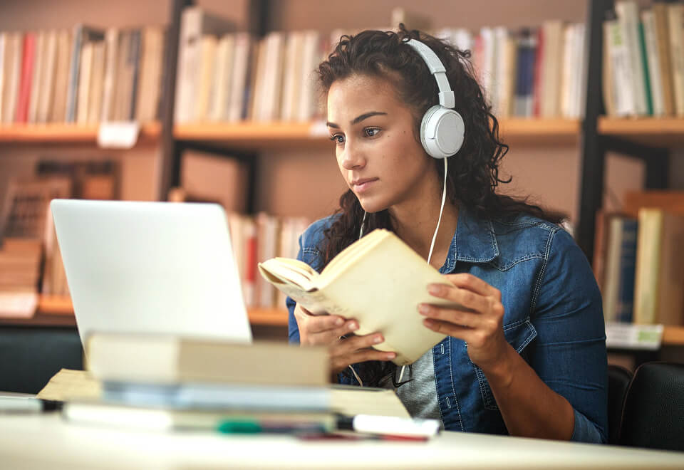 A woman is in a library listening to music while checking through a book and browsing Usenet on her laptop.
