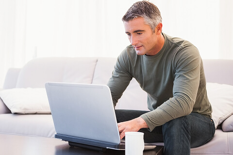 A man with salt and pepper hair is typing on a laptop. He is sitting on a white couch and has a coffee cup on the table next to him.