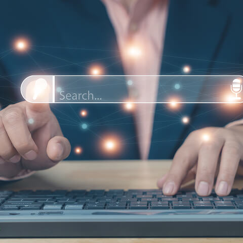 Closeup of a person’s torso. They are typing on a keyboard with one hand. There is a search box floating over the keyboard like a hologram and the person is pressing the Search button with the other hand.