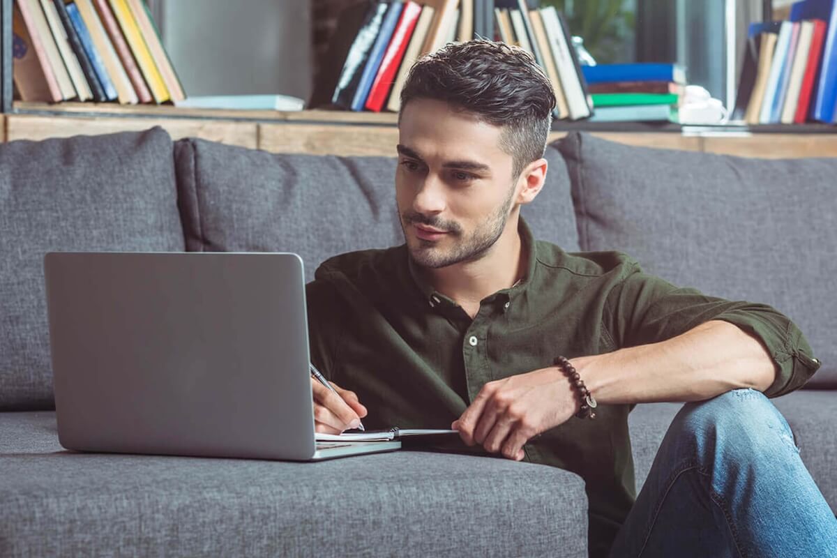 A man in a green shirt and jeans is sitting on a couch, reading a newsgroup on his laptop and taking notes in a notebook.