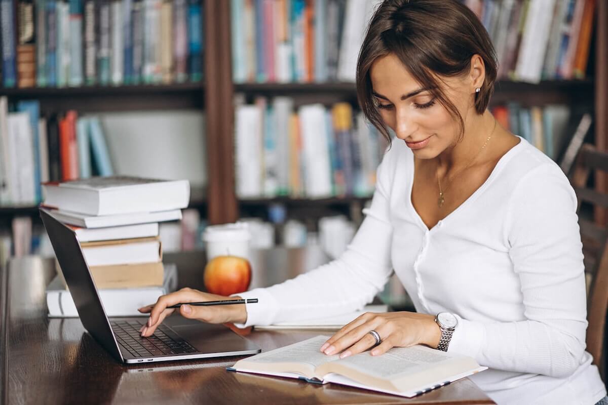 A woman in a long sleeve white shirt is looking up something in a book and typing it into a laptop, sharing that knowledge with a newsgroup.