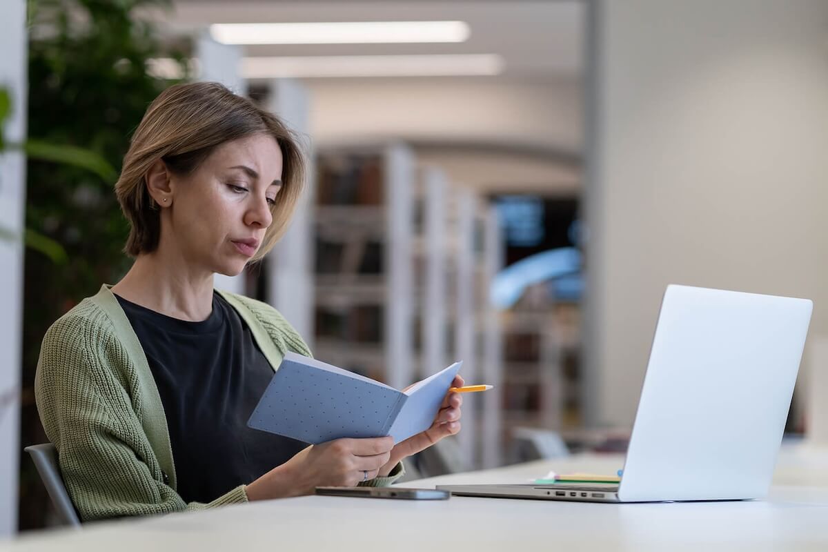 A woman wearing a green cardigan and black shirt is checking her notes in a notebook before composing a post to one of her newsgroups on the laptop in front of her.