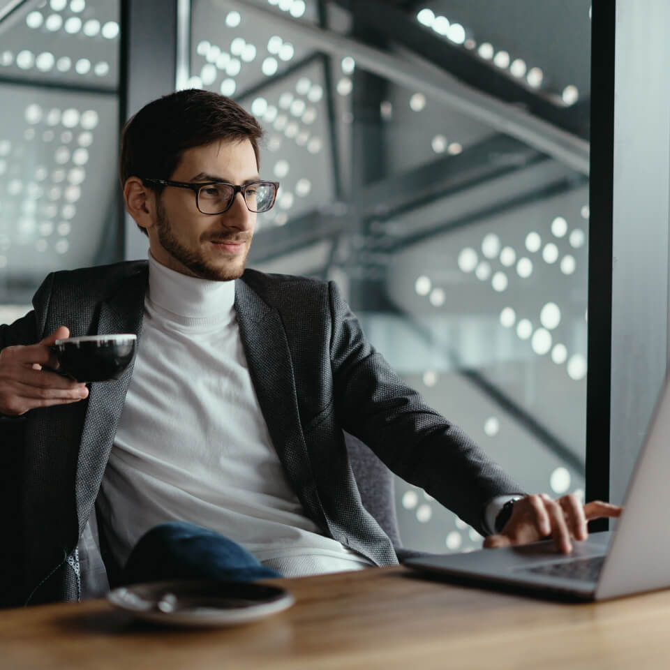 A man wearing a white turtleneck and sport coat is sitting at a conference table holding a black tea cup in his right hand while typing with his left hand on a laptop.