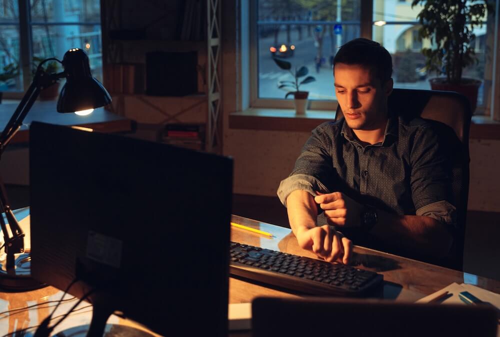 A man in a long-sleeved shirt is sitting in front of a black desktop computer and rolling up his right sleeve.