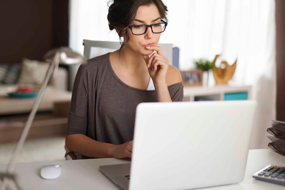 A woman wearing an off-the-shoulder grey shirt and black-framed glasses is looking at her laptop curiously, one finger under her mouth as if she’s contemplating which of the 120,000+ newsgroups she can access with Newshosting she should explore.