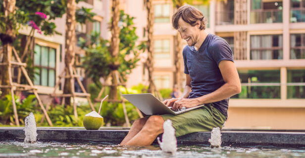 A man is sitting on the edge of a fountain with his feet in the water. He is typing on a laptop and drinking something out of a coconut with a straw stuck in it.