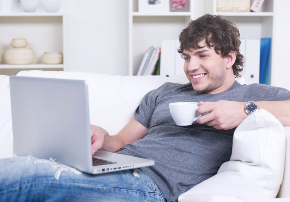 A man with short, brown hair is reclining on a couch while wearing a grey t-shirt and ripped jeans. He is holding a coffee cup in his left hand and typing on a laptop with his right hand. He is learning what Usenet is.