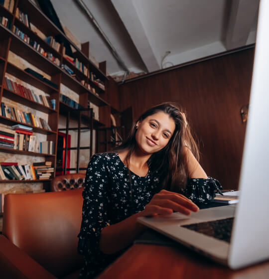 A woman is smiling and brushing her hair with her left hand while scrolling through newsgroups on a laptop with her right hand.