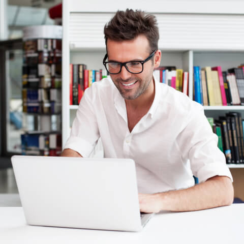 A man in a white polo shirt is in a modern library and smiling as he works on his laptop.